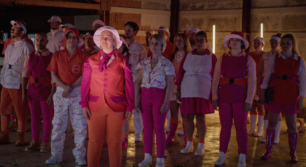 Maren Montalbano in the foreground looking into the distance, with a choir behind her. Everyone is wearing pink and orange costumes depicting farm or office workers.