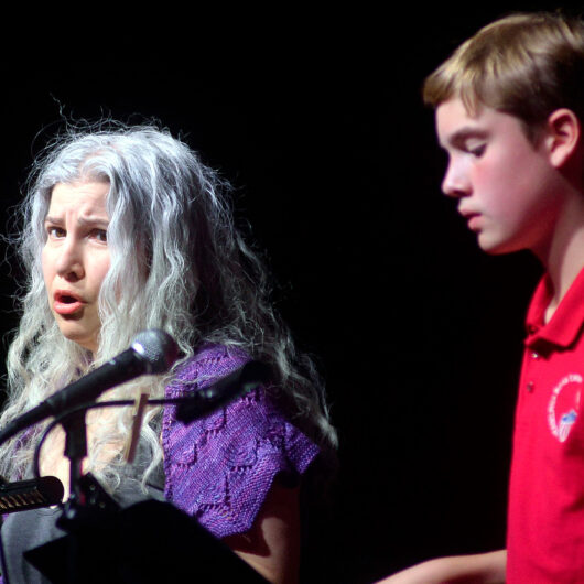 Maren Montalbano, playing The Mother, singing into a microphone, standing next to a young boy, Ethan Monberg, who plays Amahl.